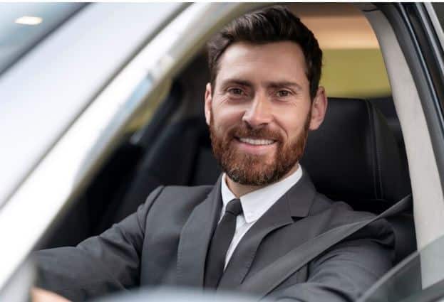 A professional chauffeur in a formal suit and tie stands next to a sleek, black luxury sedan, ready to provide transportation services- ambassador chauffeur